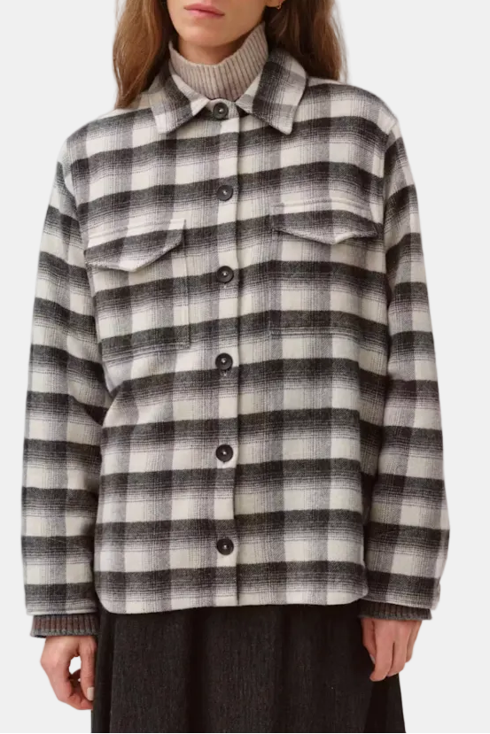 A woman wearing a black and off-white checkered button-up shirt jacket over a ribbed beige turtleneck, photographed against a plain light gray studio background.