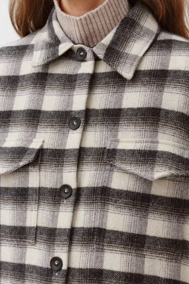 Close-up of a person wearing a black and off-white plaid flannel shirt over a beige ribbed turtleneck, with the collar and buttons visible.