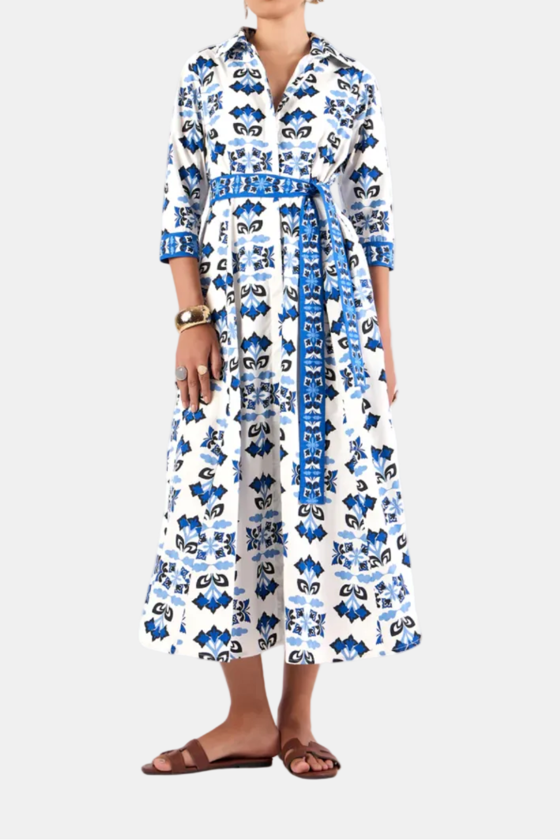 A woman standing against a white background wearing a long blue and white floral dress with 3/4 length sleeves.