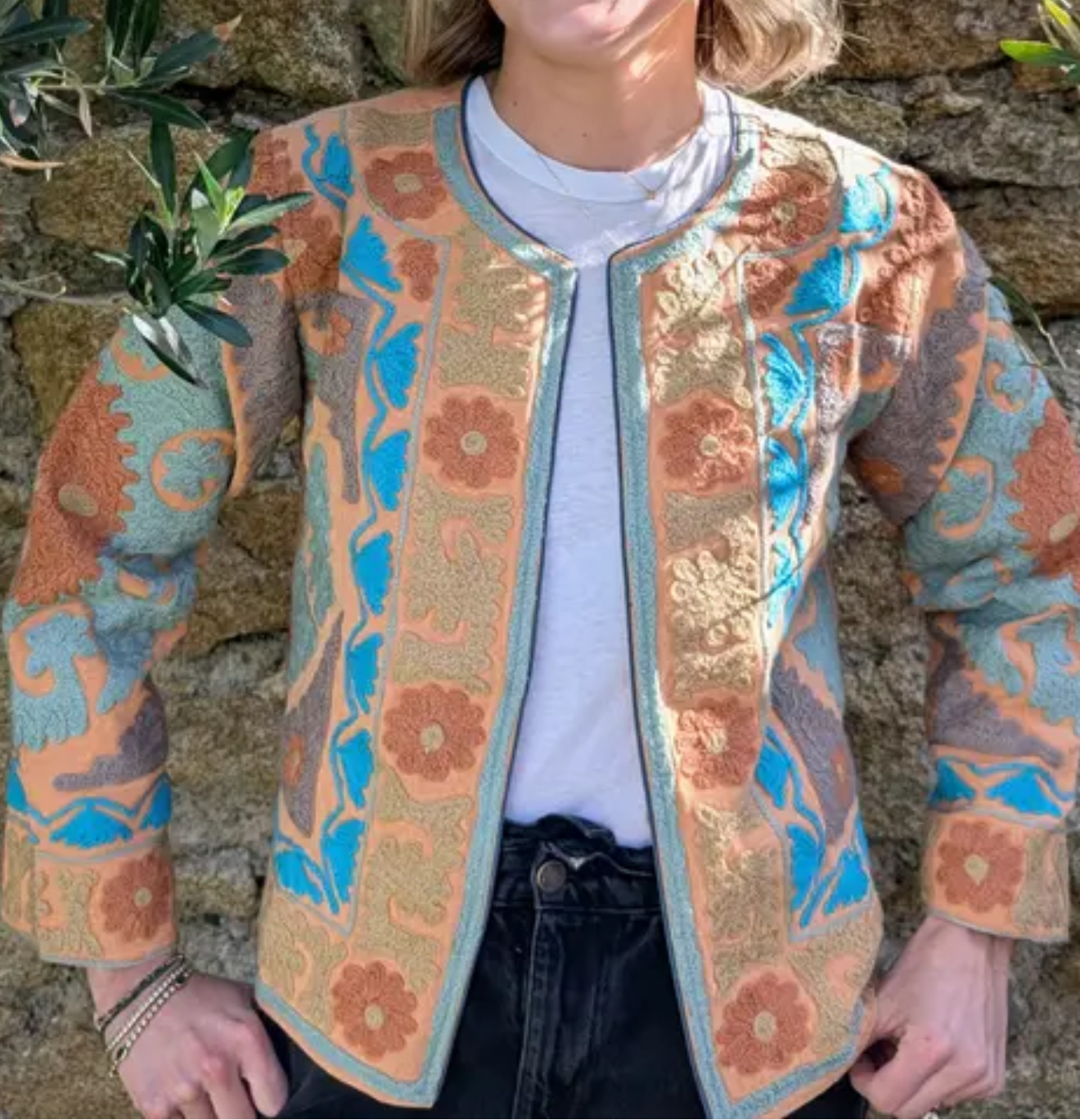Woman wearing a patterned jacket and sunglasses against a stone wall.