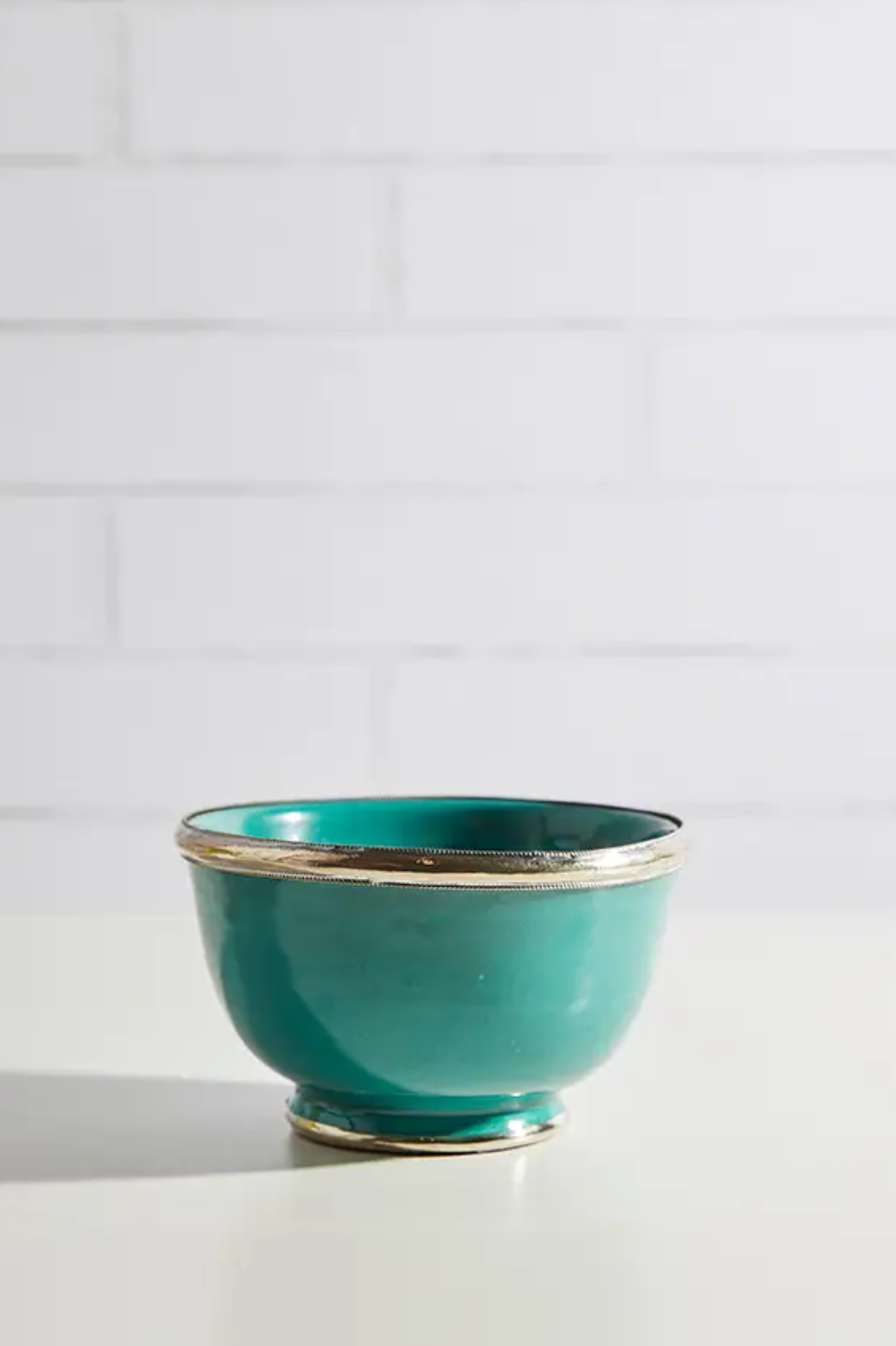 Teal ceramic bowl with gold rim on a white surface with a light gray tiled wall background