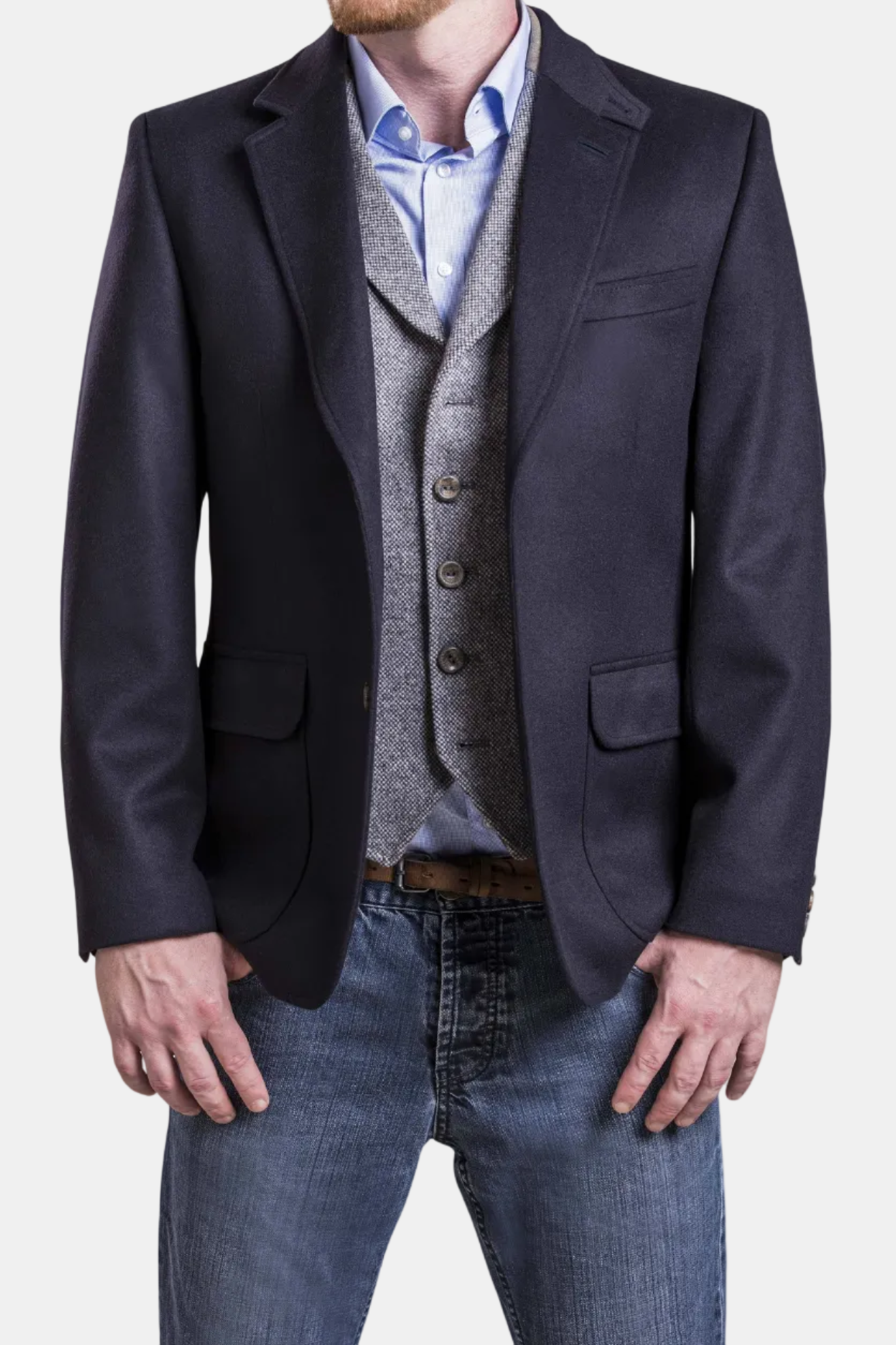 Man wearing a dark blazer, light blue shirt, gray vest, and jeans on a white background