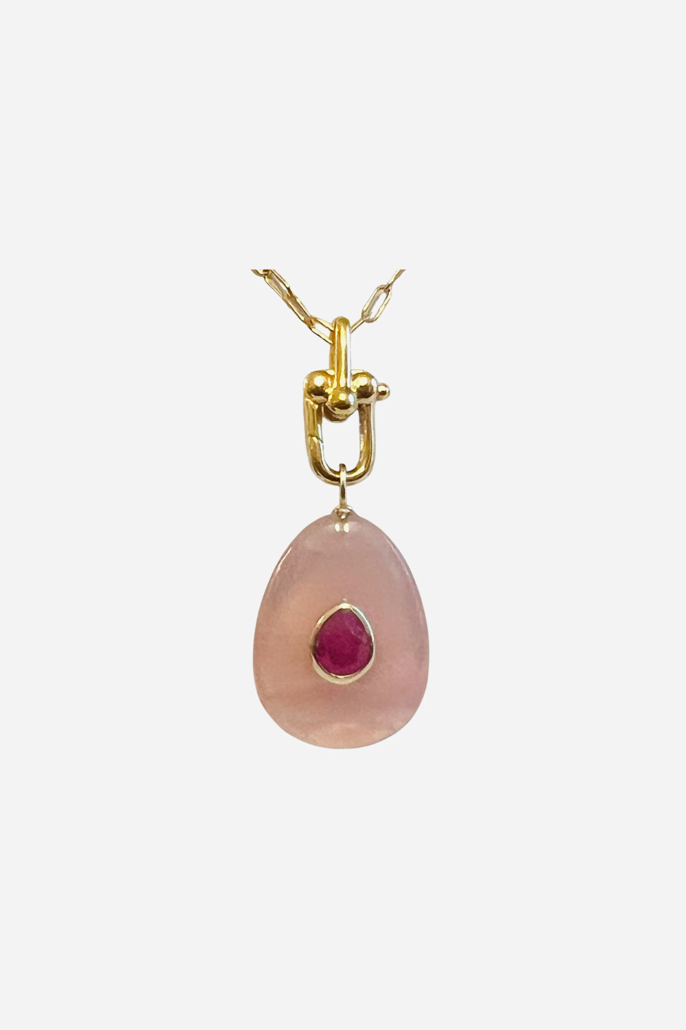 A gold pendant necklace featuring a teardrop-shaped light pink stone with a small, round, deep pink gemstone set in the center, suspended from a delicate gold chain against a plain light gray background.