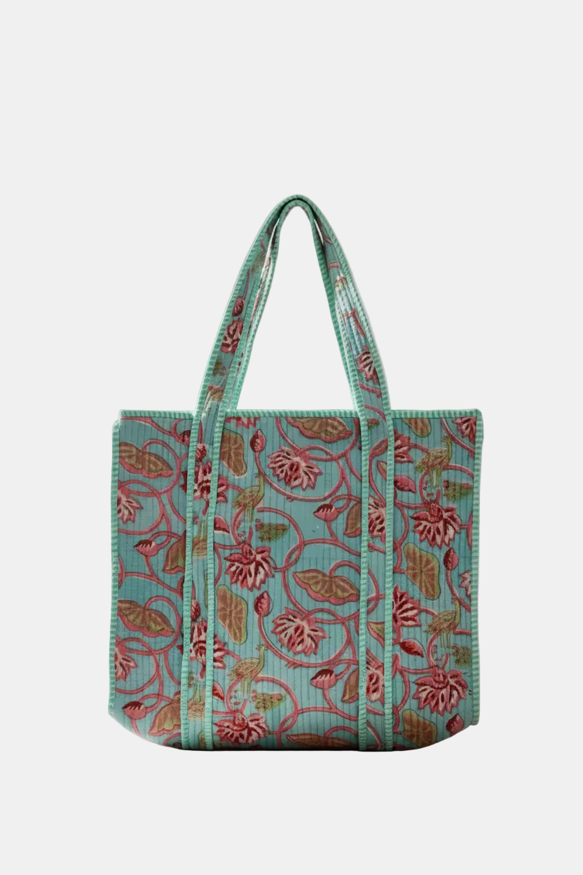 A structured tote bag with a turquoise blue background and a pink and green floral vine pattern, displayed on a light wood table in a bright, modern living room setting.