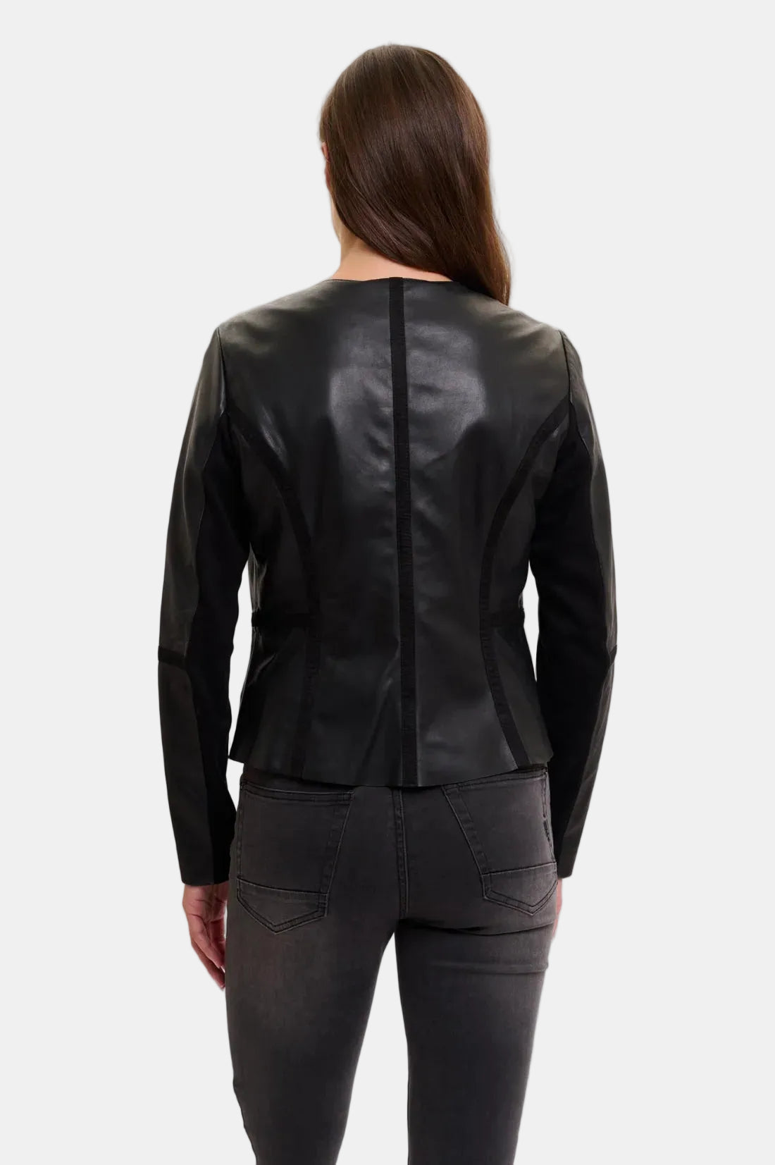 A woman viewed from the back wearing a fitted black leather jacket and dark gray jeans against a plain light gray studio background.