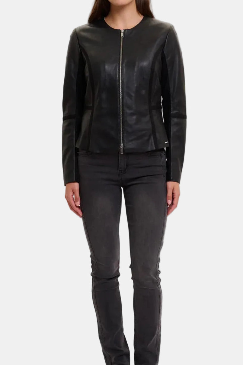 A woman modeling a black leather jacket with a front zipper, paired with dark gray skinny jeans and black loafers, against a plain light gray studio background.