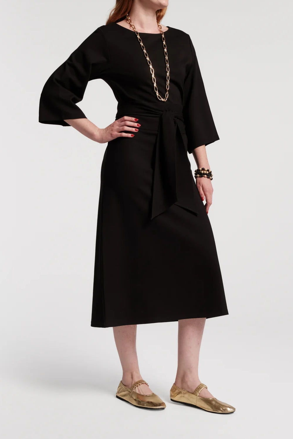 A woman modeling a black midi dress with three-quarter sleeves, a round neckline, and a self-tie waist, styled with a chunky gold chain necklace and gold flat shoes against a light gray studio background.