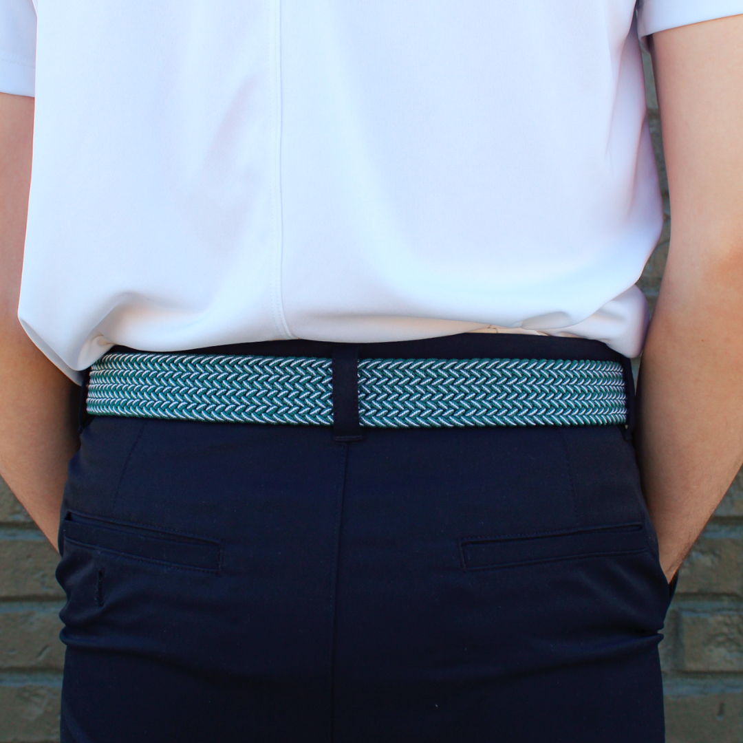 A close-up back view of a person wearing a white short-sleeve shirt, dark navy trousers, and a woven belt with a black and light blue chevron pattern, standing against a brick wall.