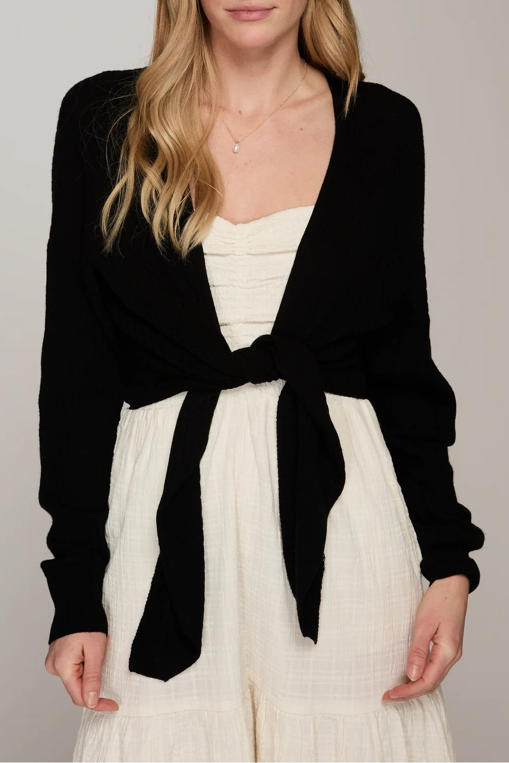 Woman wearing a black cardigan over a white top and pants on a plain background