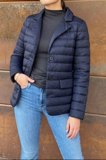 Woman wearing a navy puffer jacket and blue jeans standing against a rusty metal wall.