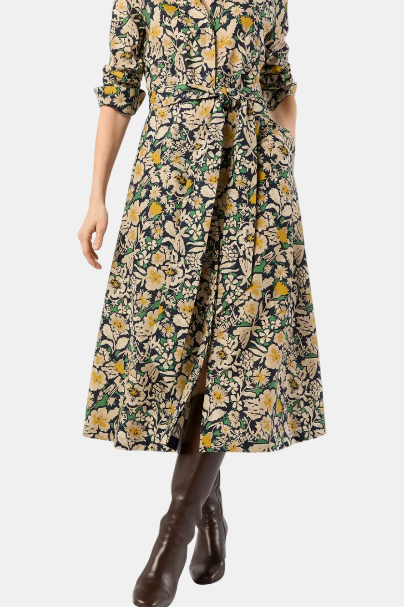 A woman wearing a long-sleeved midi wrap dress with a dense floral and botanical print in green, beige, and yellow, paired with dark brown leather ankle boots, shown against a plain white background.