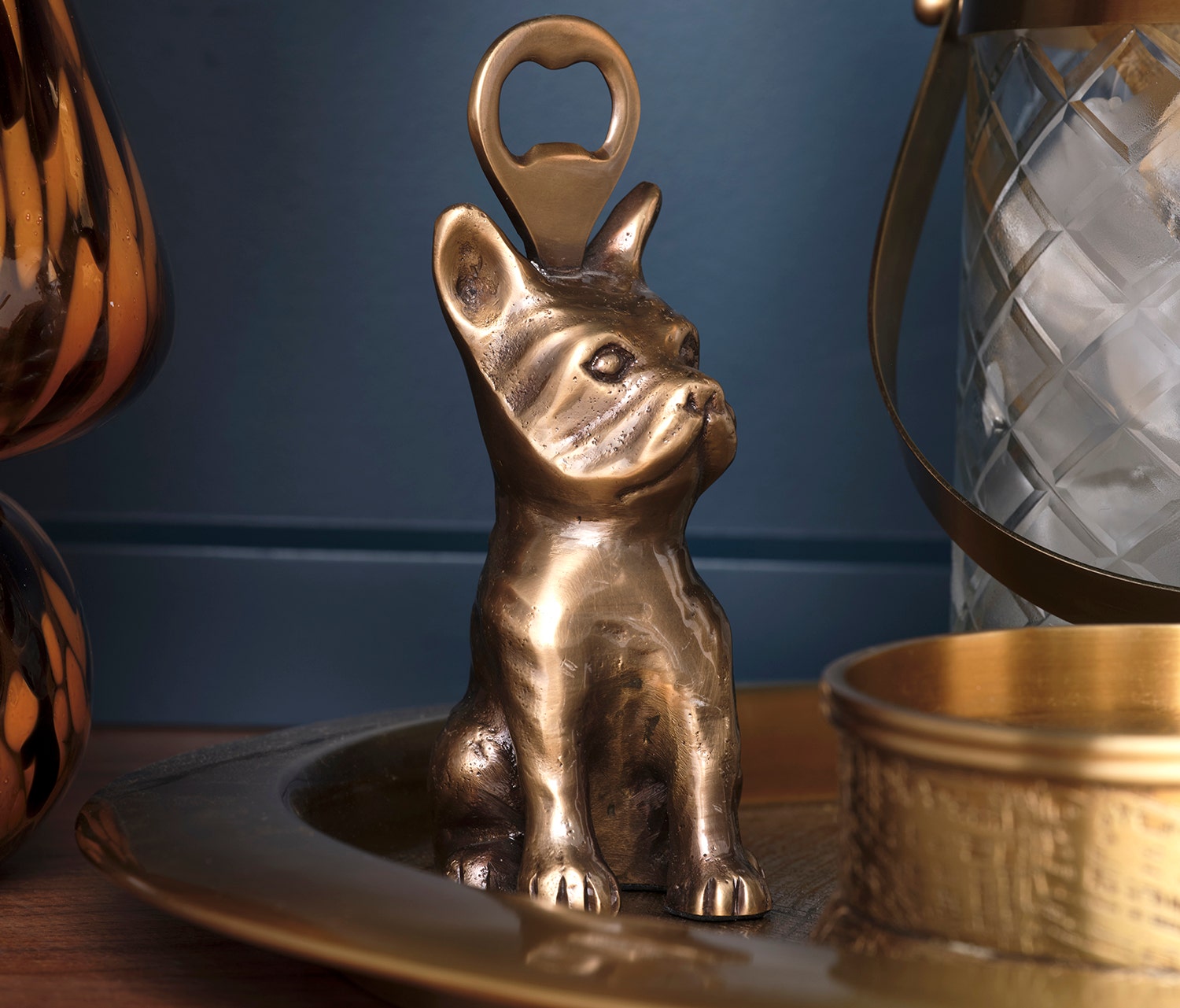 A brass French Bulldog-shaped bottle opener resting on a matching brass tray, with a blue paneled background and decorative home decor items partially visible.