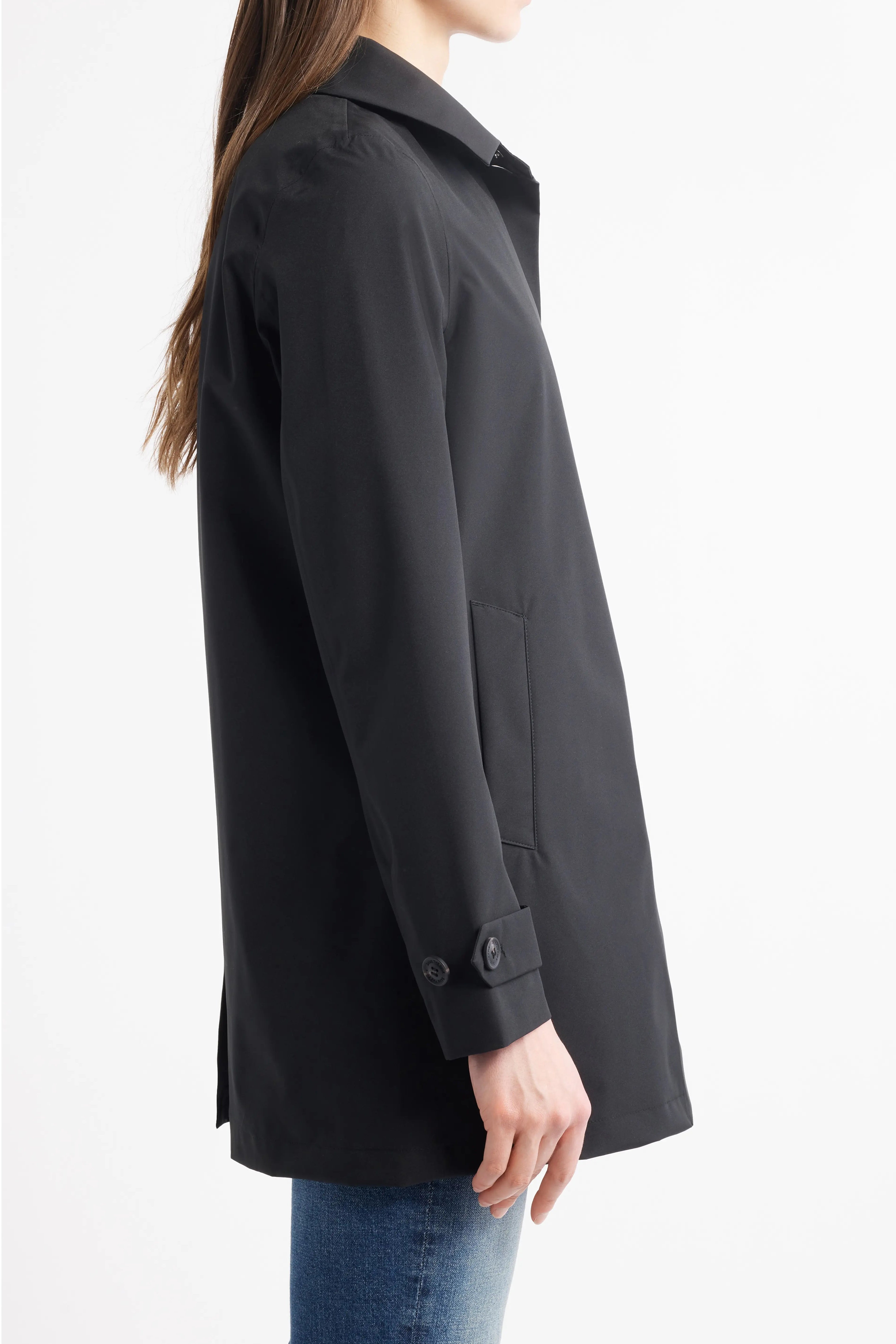 A woman shown from the side wearing a black mid-length coat with a collar, buttoned cuffs, and front patch pockets, paired with blue jeans against a plain white studio background.