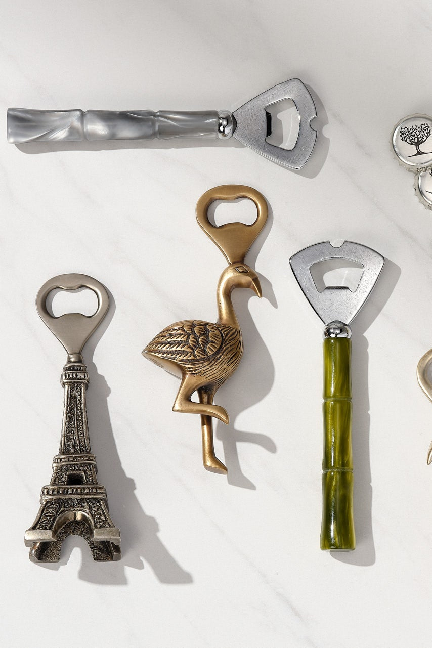 A set of decorative bottle openers arranged on a white marble surface, including Eiffel Tower and flamingo-shaped designs with metallic silver, gold, and green handles.