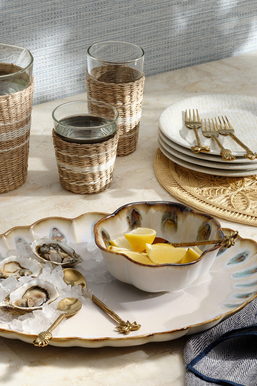 A styled dining table setting featuring a large scalloped white ceramic platter with gold trim, a matching bowl with butter, glassware wrapped in woven straw, stacked white plates with gold forks, and a gray napkin on a light marble surface.