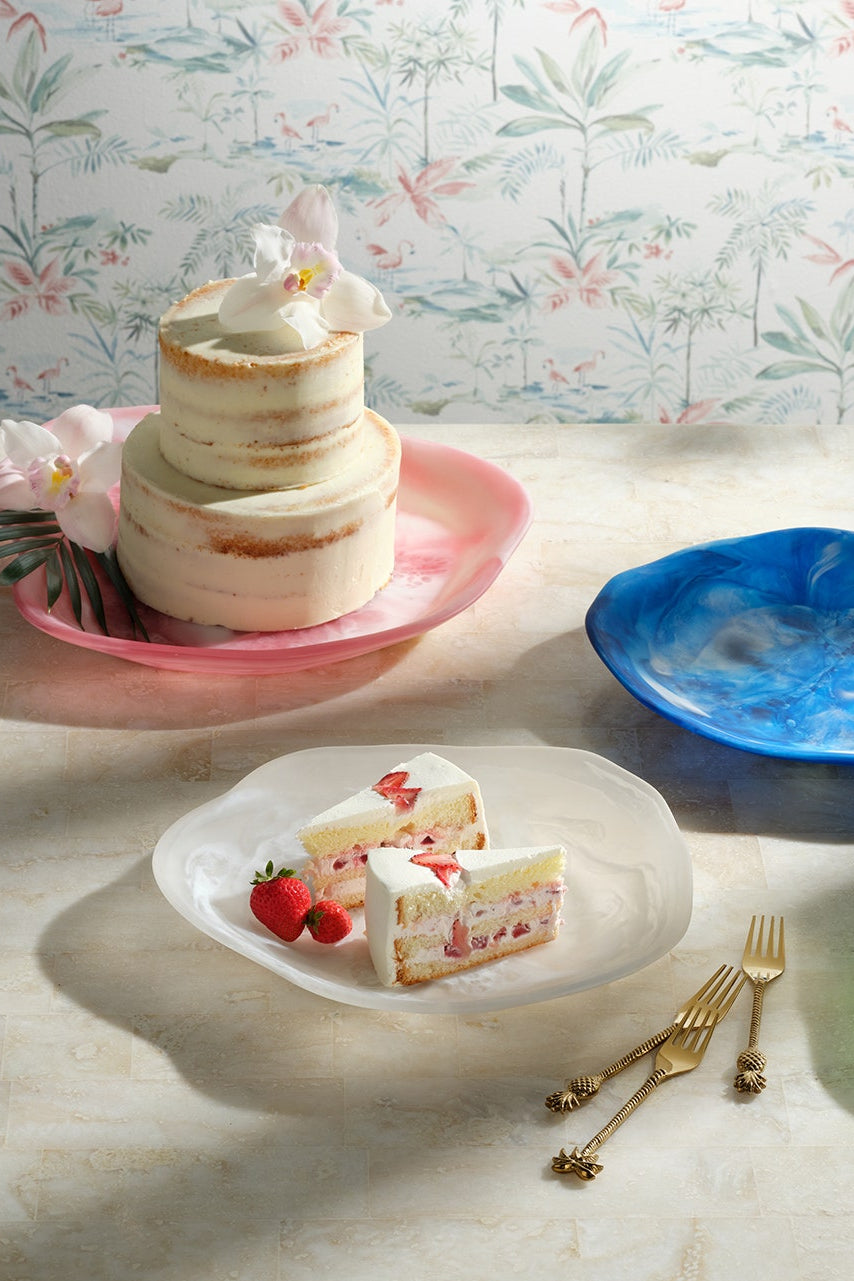 A styled tabletop scene showing several translucent wavy-edged plates in pink, blue, green, and peach, with two small cakes and sliced cake on some plates, set against floral wallpaper and a light stone surface.