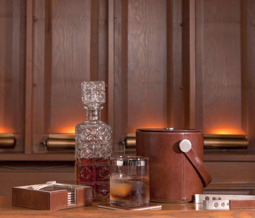 A whiskey decanter, a leather-look ice bucket with silver handle, a clear drinking glass with amber liquid, and a small leather box arranged on a wooden surface against a warm wood-paneled background.
