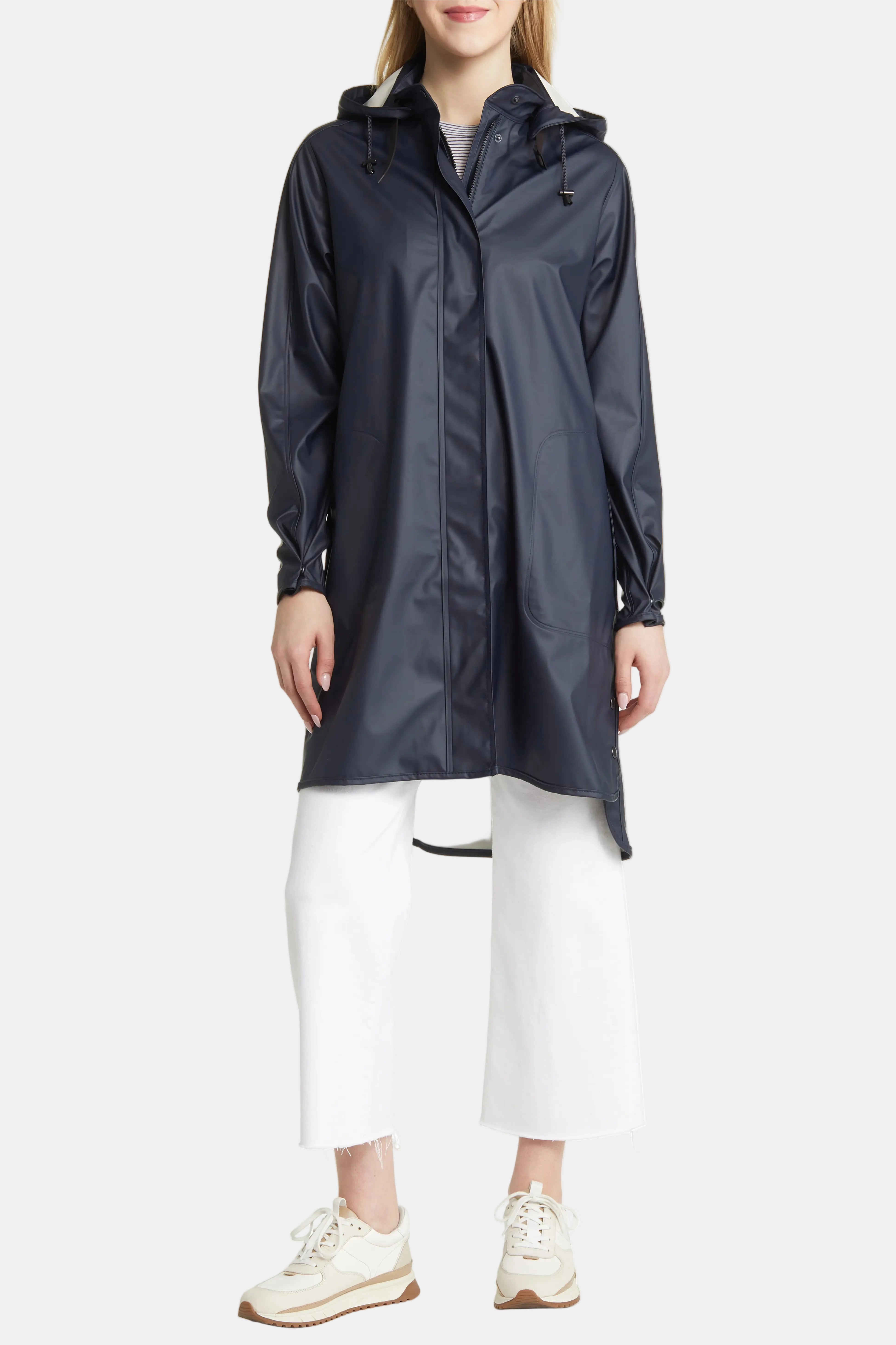 A woman wearing a dark navy hooded raincoat over a gray top and white cropped pants, standing against a plain white studio background.