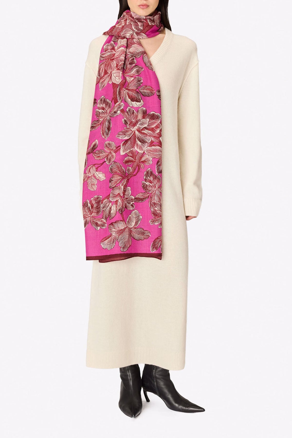 A model wearing a long cream-colored coat and black pointed boots, wrapped in a vibrant pink scarf with a brown and gold floral pattern, against a plain white background.