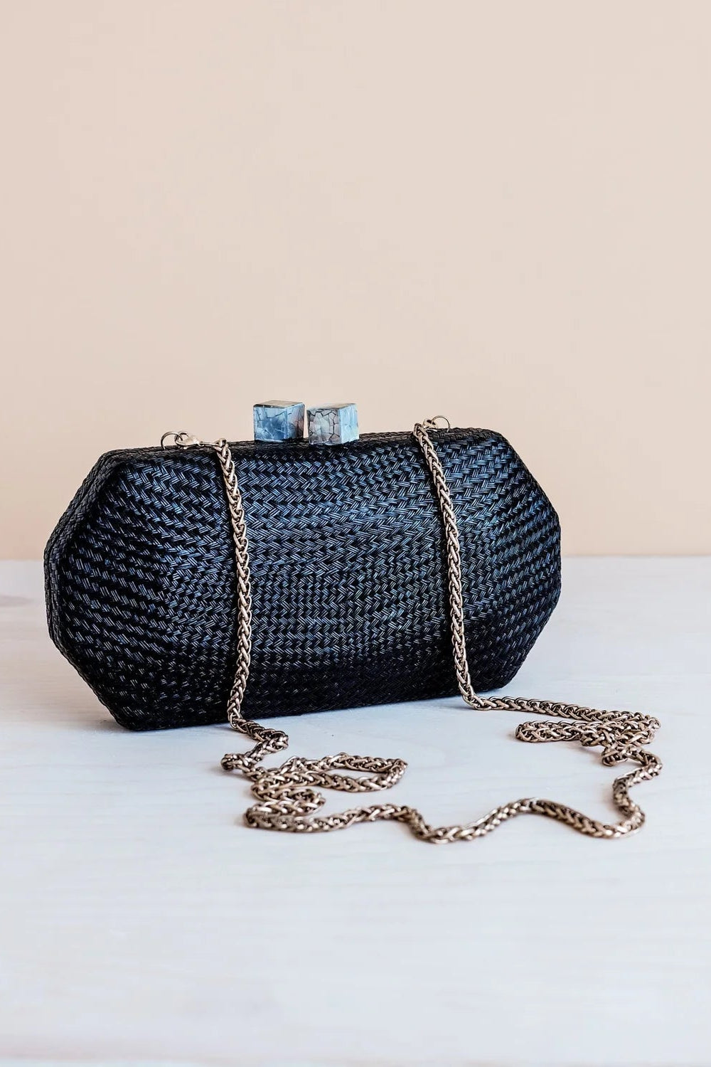 A black woven clutch bag with a gold chain strap and two blue stone clasp details, resting on a white surface against a beige background.