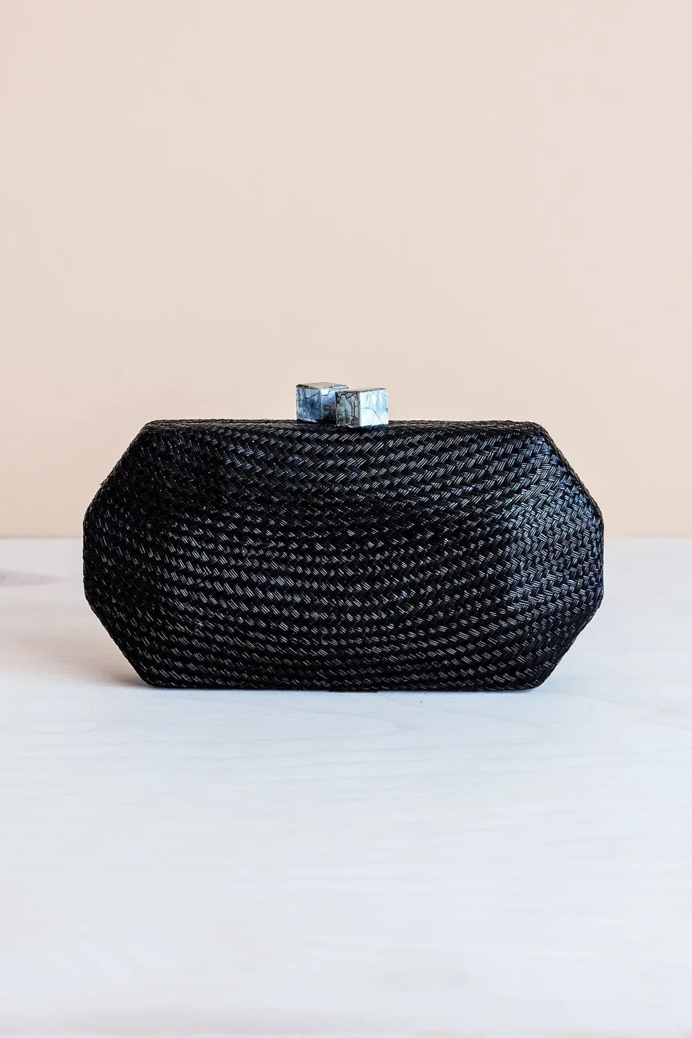 A small black woven clutch bag with a geometric hexagonal shape and a silver-toned metal clasp, resting on a white surface against a light beige background.