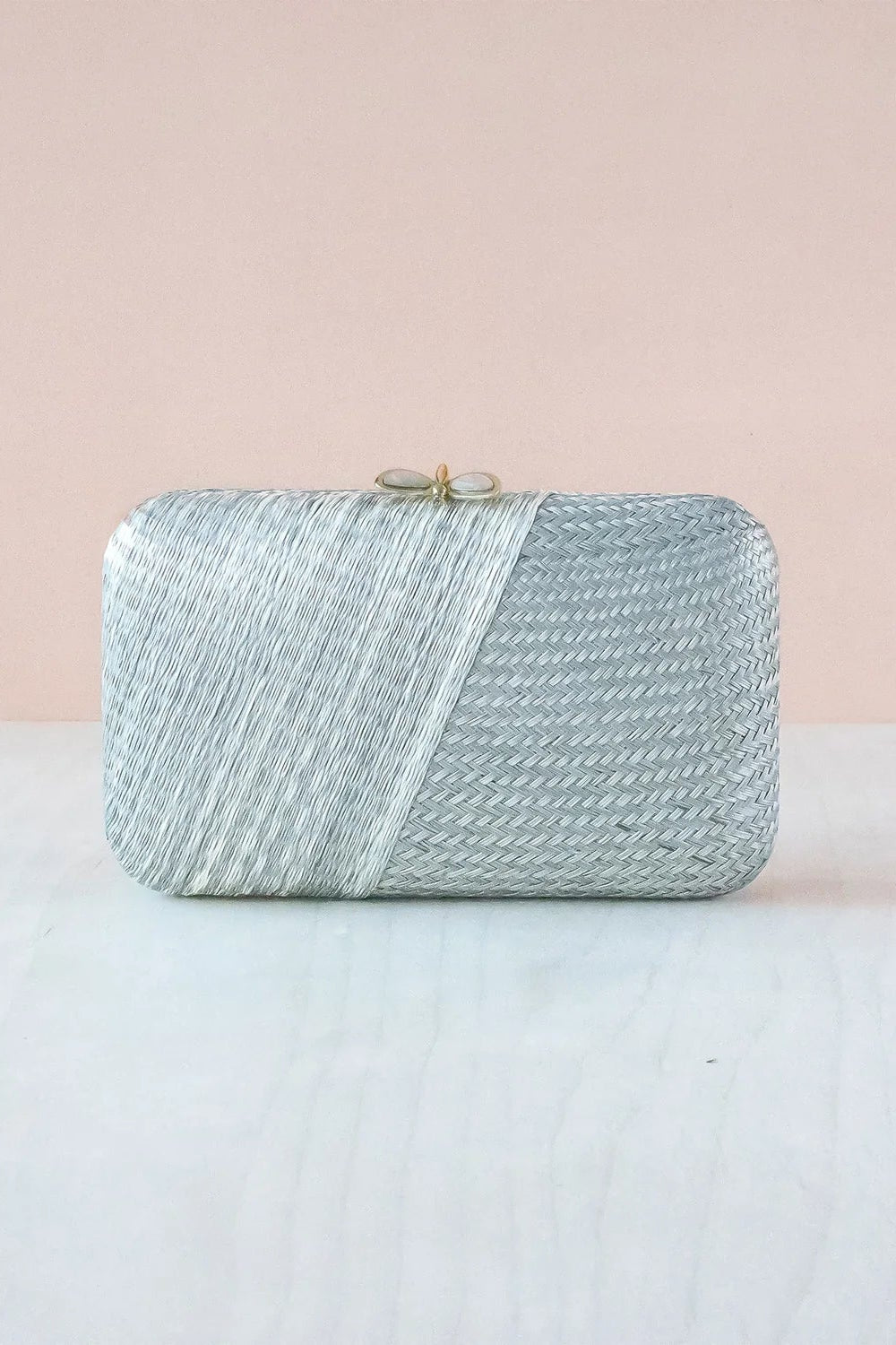 A silver metallic woven clutch purse with a diagonal split design and a small gold clasp, resting on a light surface against a soft pink background.