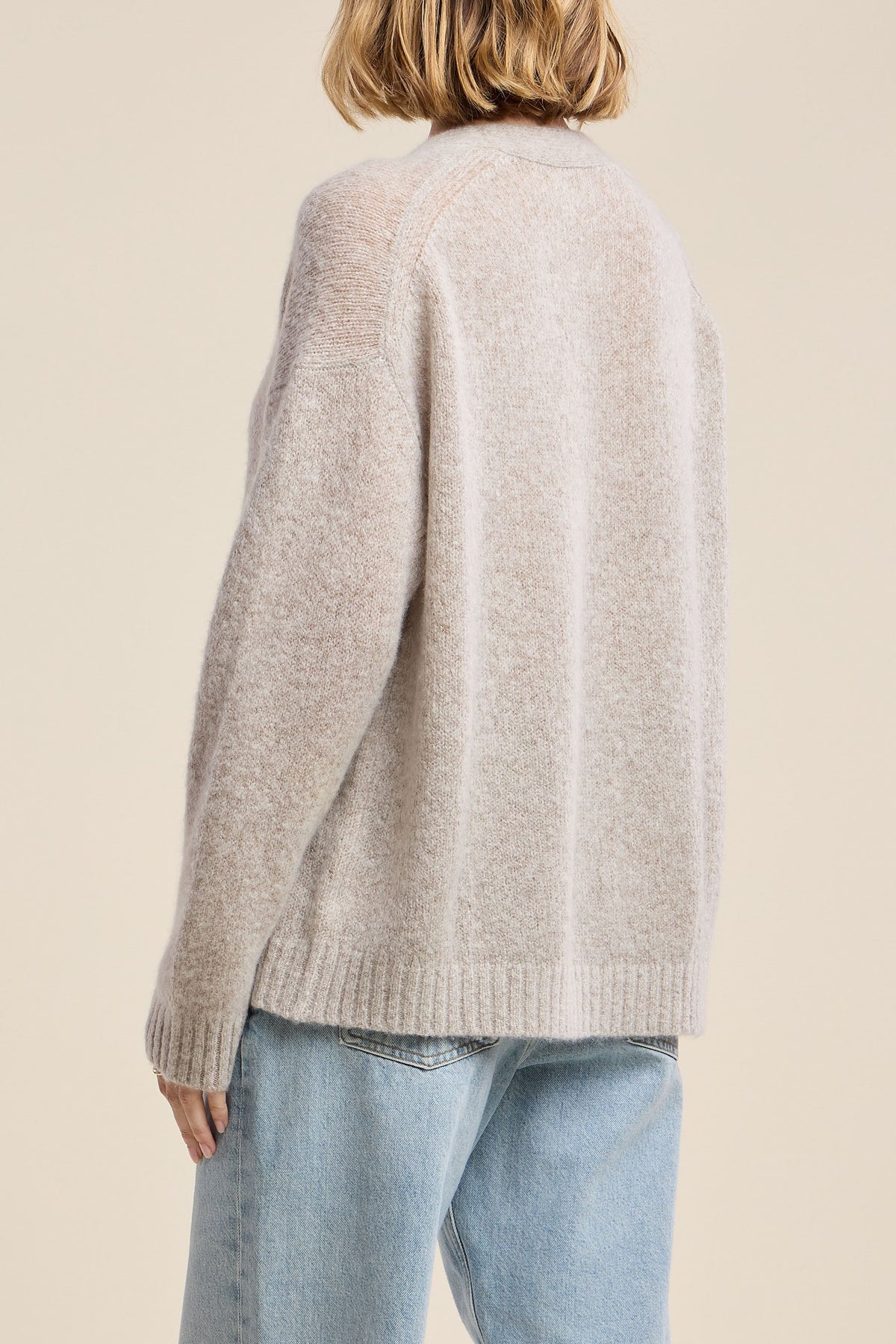 A woman shown from the back wearing a light beige knit sweater and light blue jeans, set against a solid warm beige studio background.