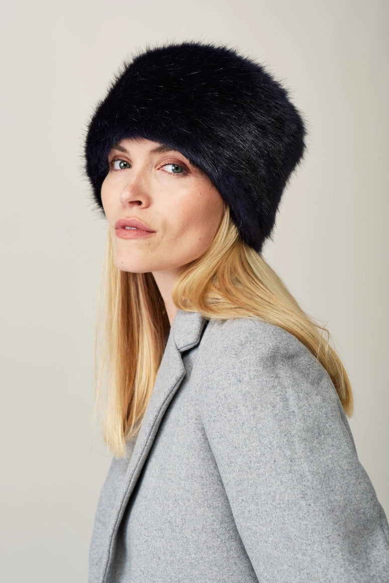 A blonde woman wearing a black faux fur ushanka hat and a gray blazer, photographed against a neutral light gray background.