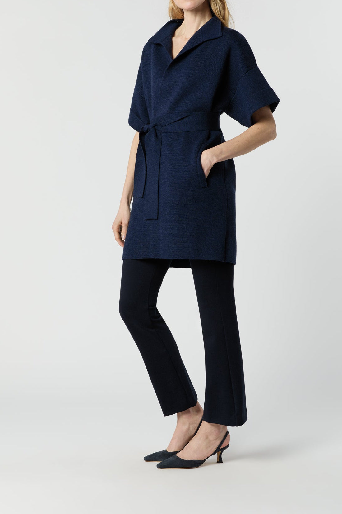 A woman modeling a dark navy blue belted wrap coat with short sleeves and a collar, paired with dark trousers and black heels, against a light gray studio background.