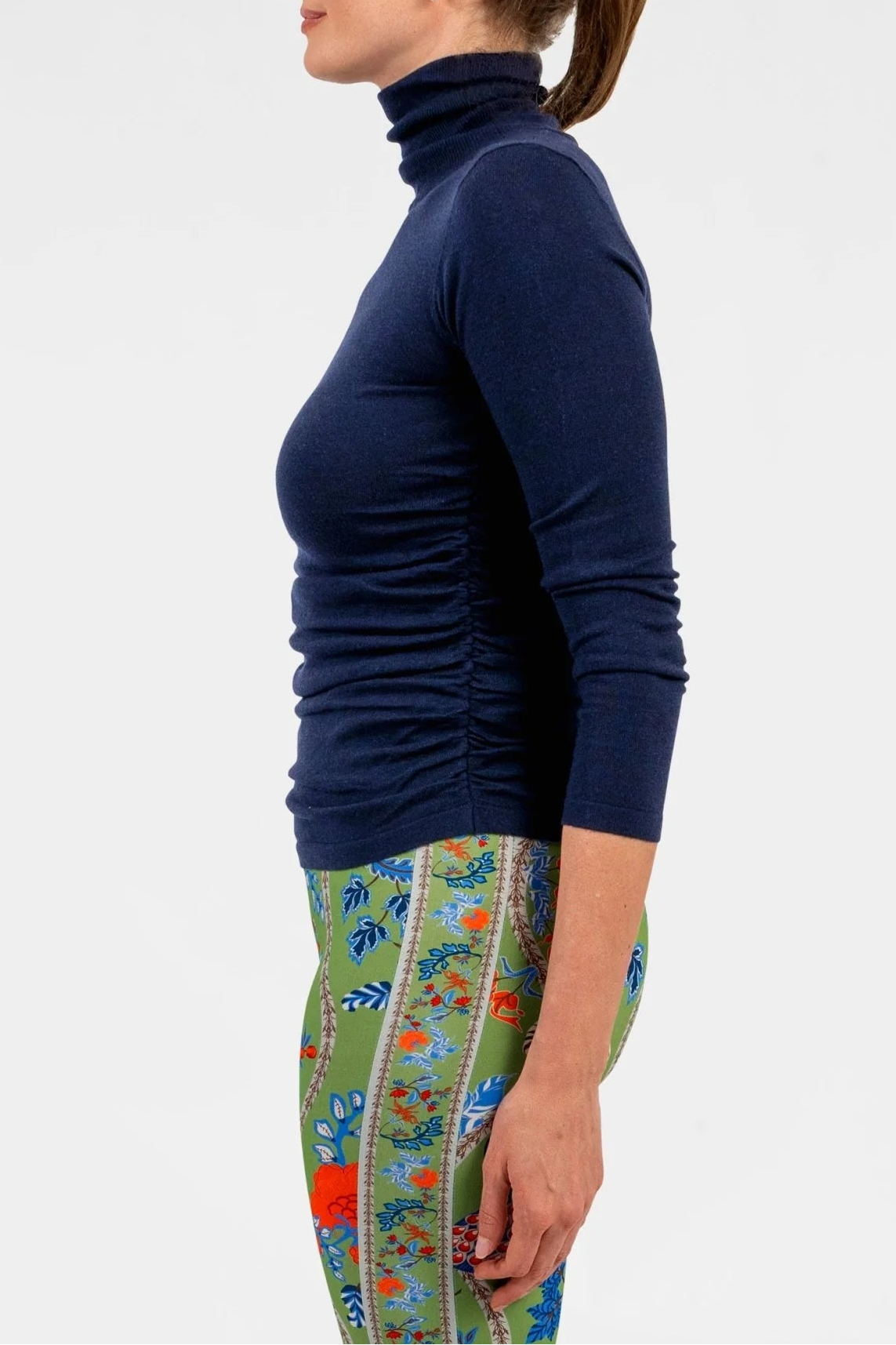 A woman shown in profile wearing a fitted navy blue long-sleeve turtleneck top and colorful patterned leggings against a plain light gray studio background.