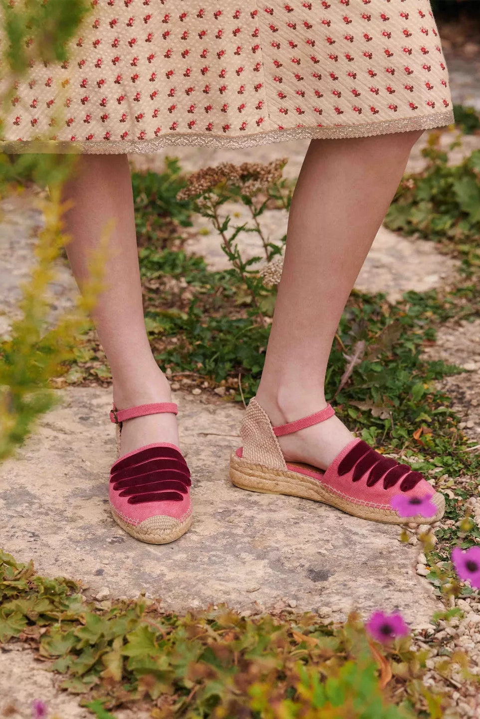 Person wearing pink sandals and a patterned skirt standing on a stone path with greenery.