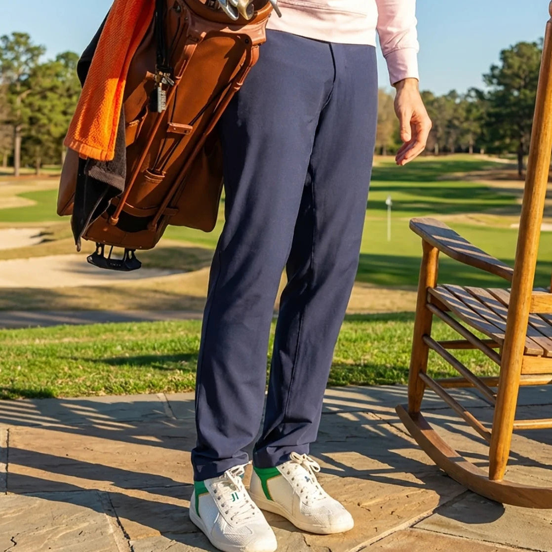 Person standing on a patio with a golf bag, wearing navy pants and white sneakers, with a wooden rocking chair and golf course in the background.