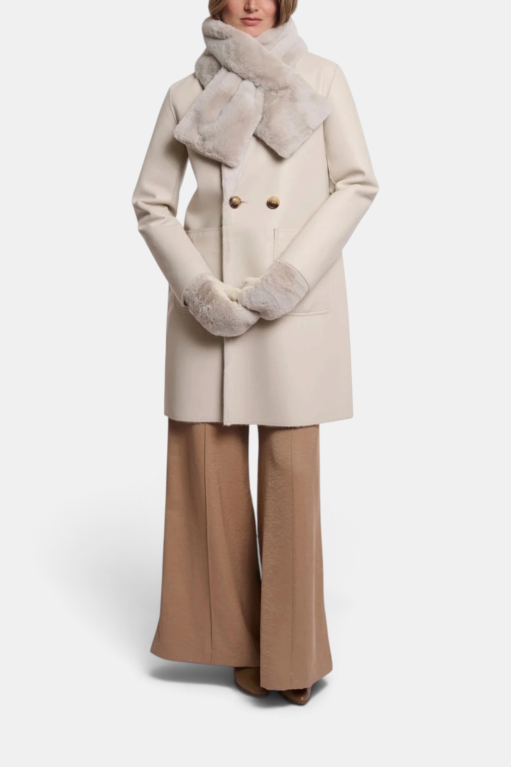 A woman modeling a double-breasted beige wool coat with a plush light beige faux-fur collar, matching faux-fur gloves, and wide-leg tan trousers, photographed against a seamless white studio background.