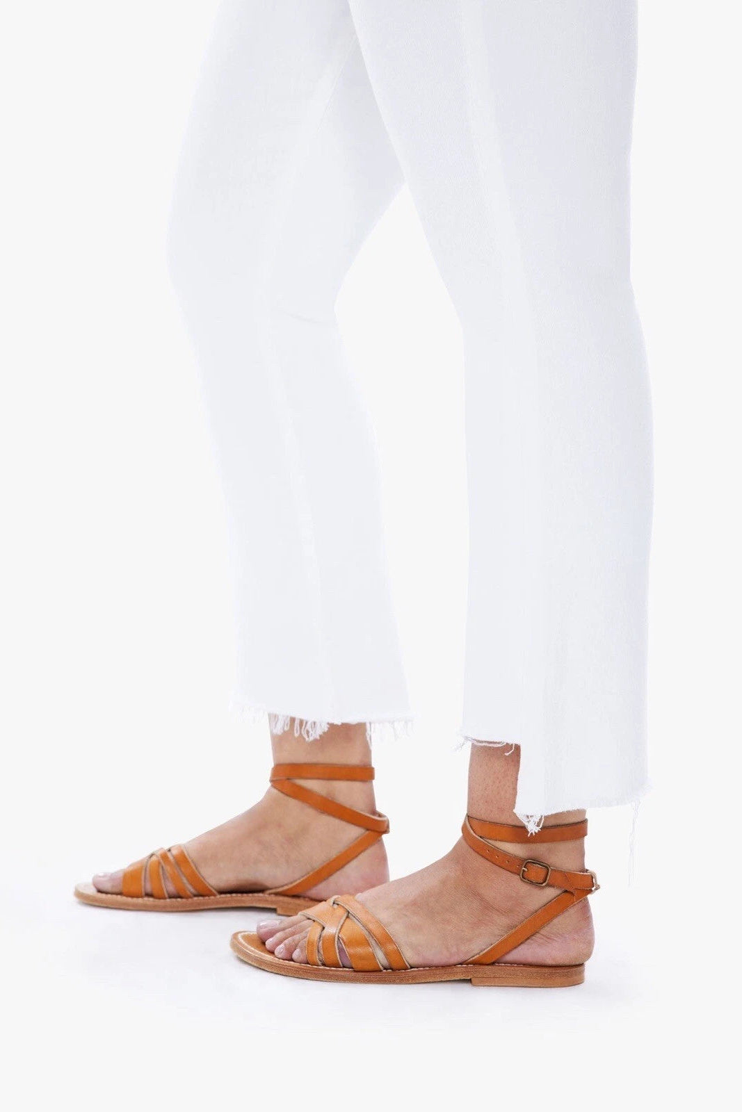 A person wearing tan leather strappy flat sandals with ankle straps, paired with white cropped jeans, shown from the calves down on a clean white background.