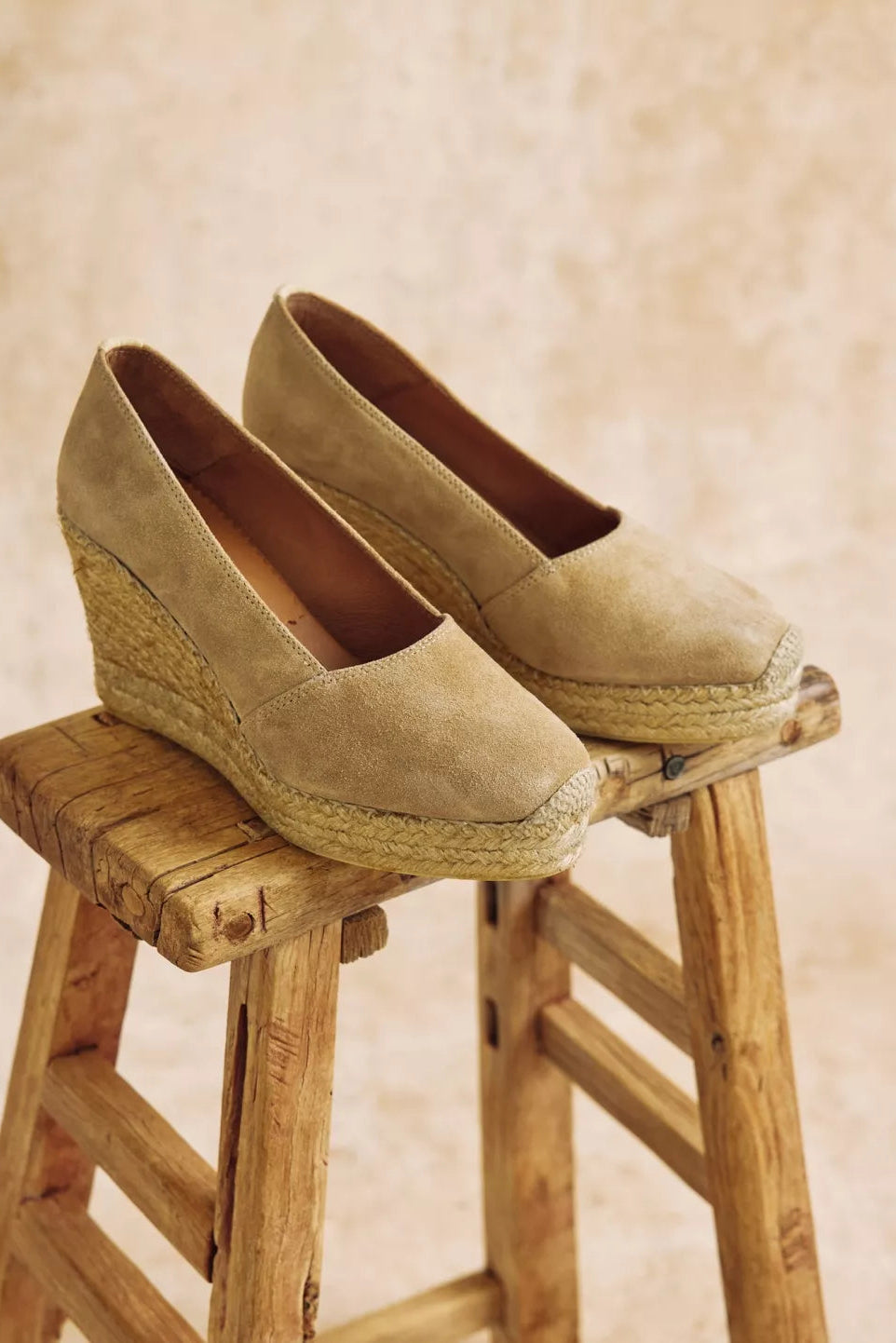 Pair of beige wedge shoes on a wooden stool with a beige background