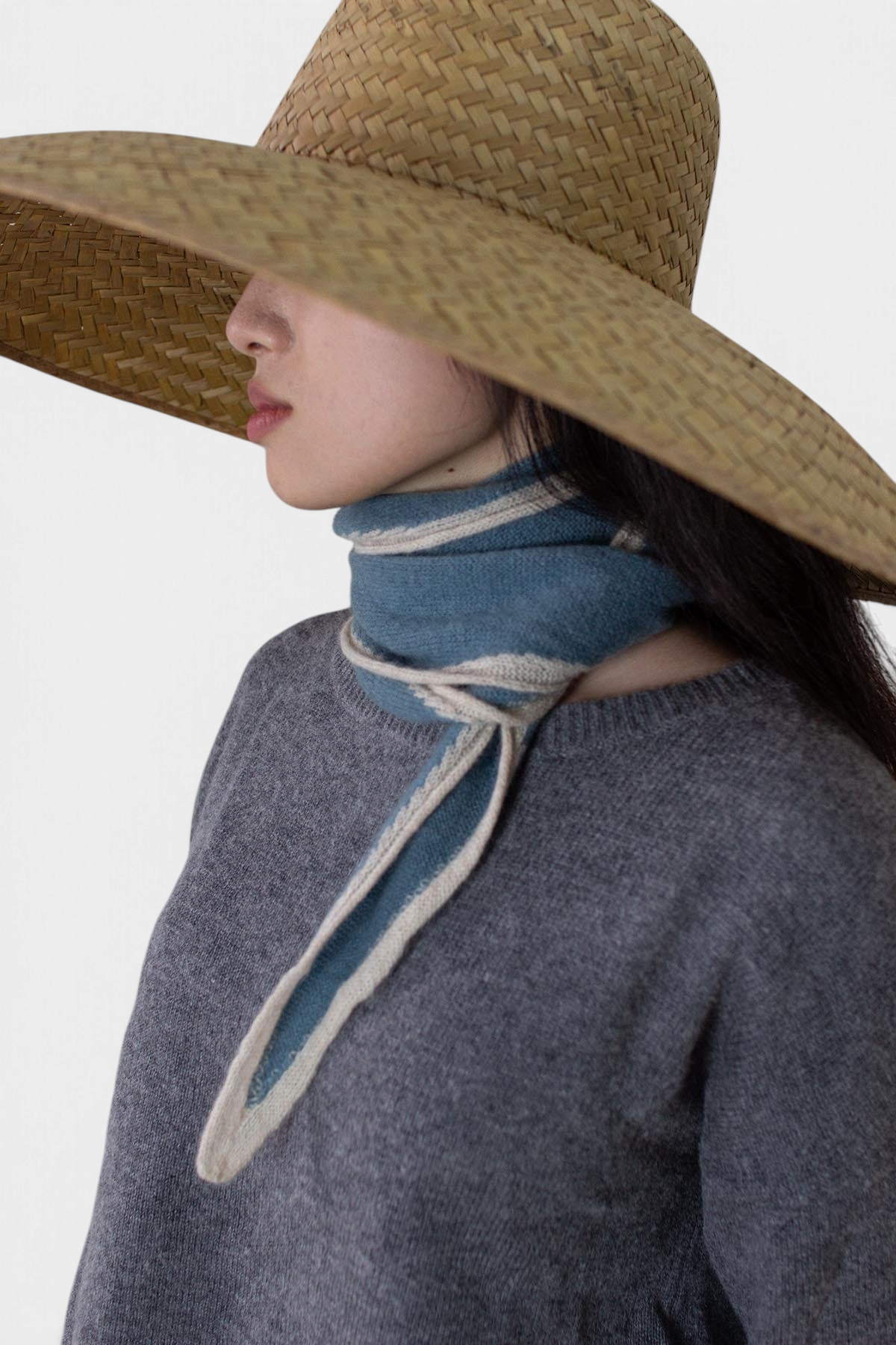 The Copper Shed Austen spindle-shaped blue and beige striped organic cashmere scarf worn by a model in a straw hat.