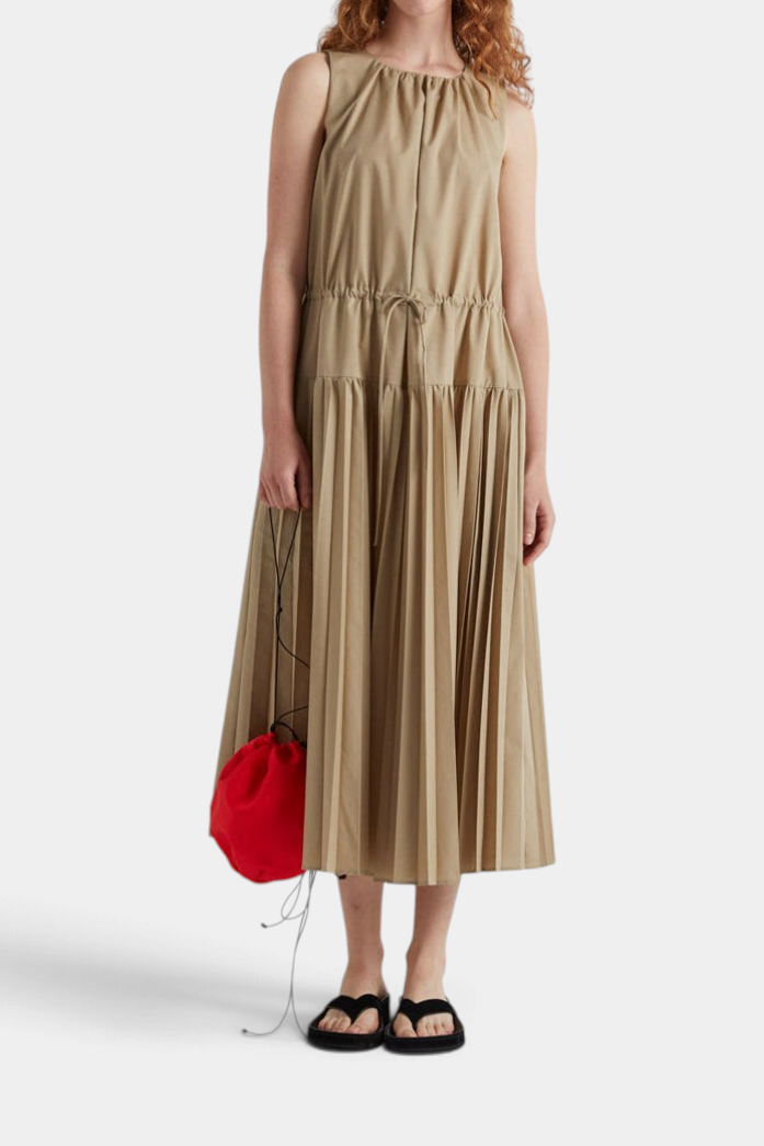 The Copper Shed long beige dress with a round neck, drawstring waist, and pleated skirt, worn with black sandals.