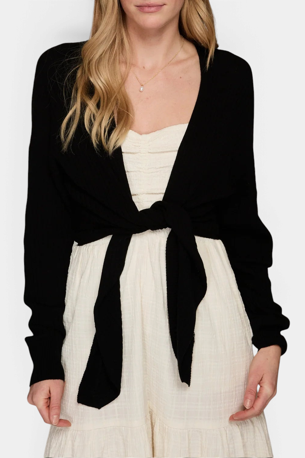 The Copper Shed black ribbed cashmere cardigan with a tie front, worn over a white dress by a model.