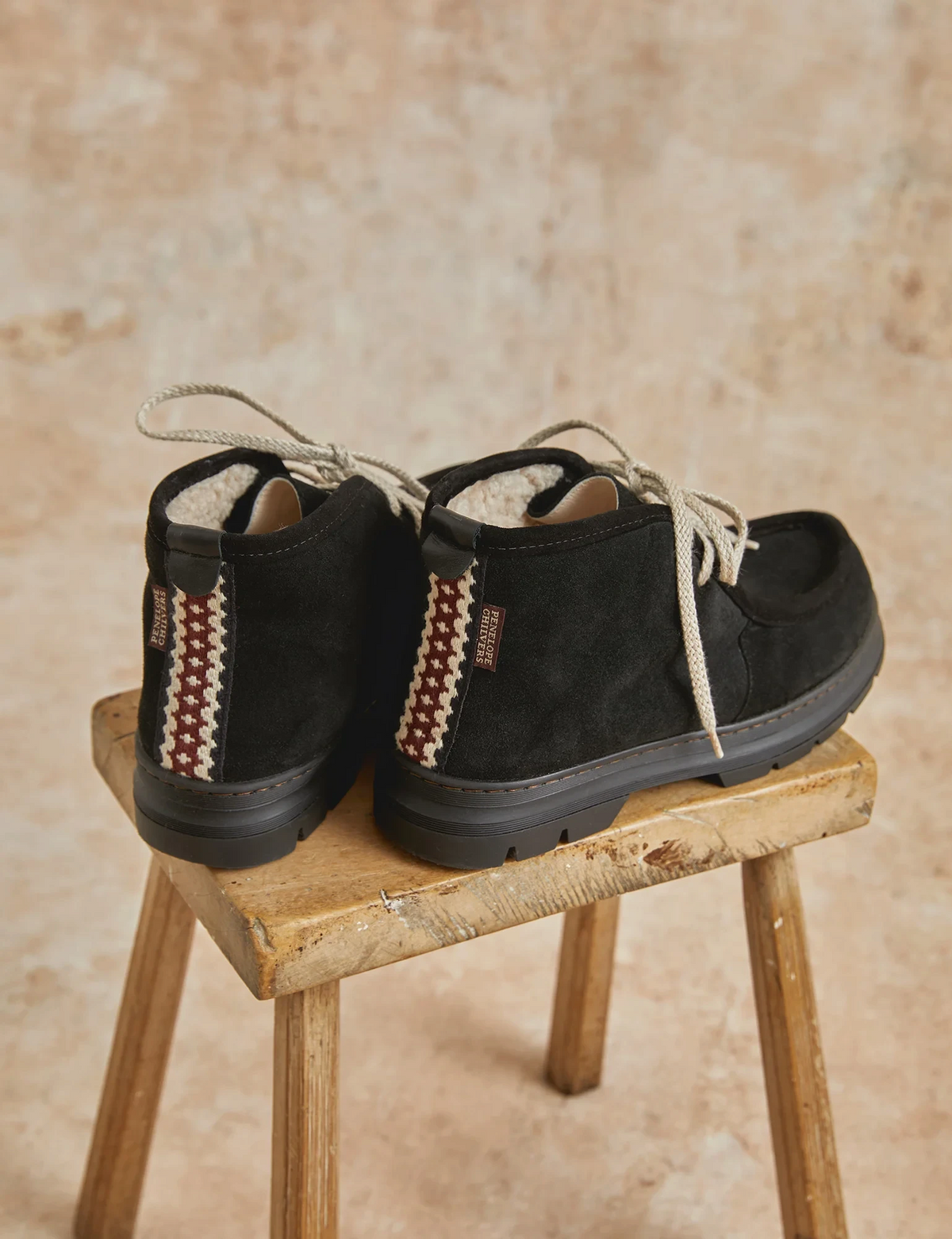 The Copper Shed Phenomenal black suede shearling-lined boots with ecru laces and folk trim heel detail on a stool.