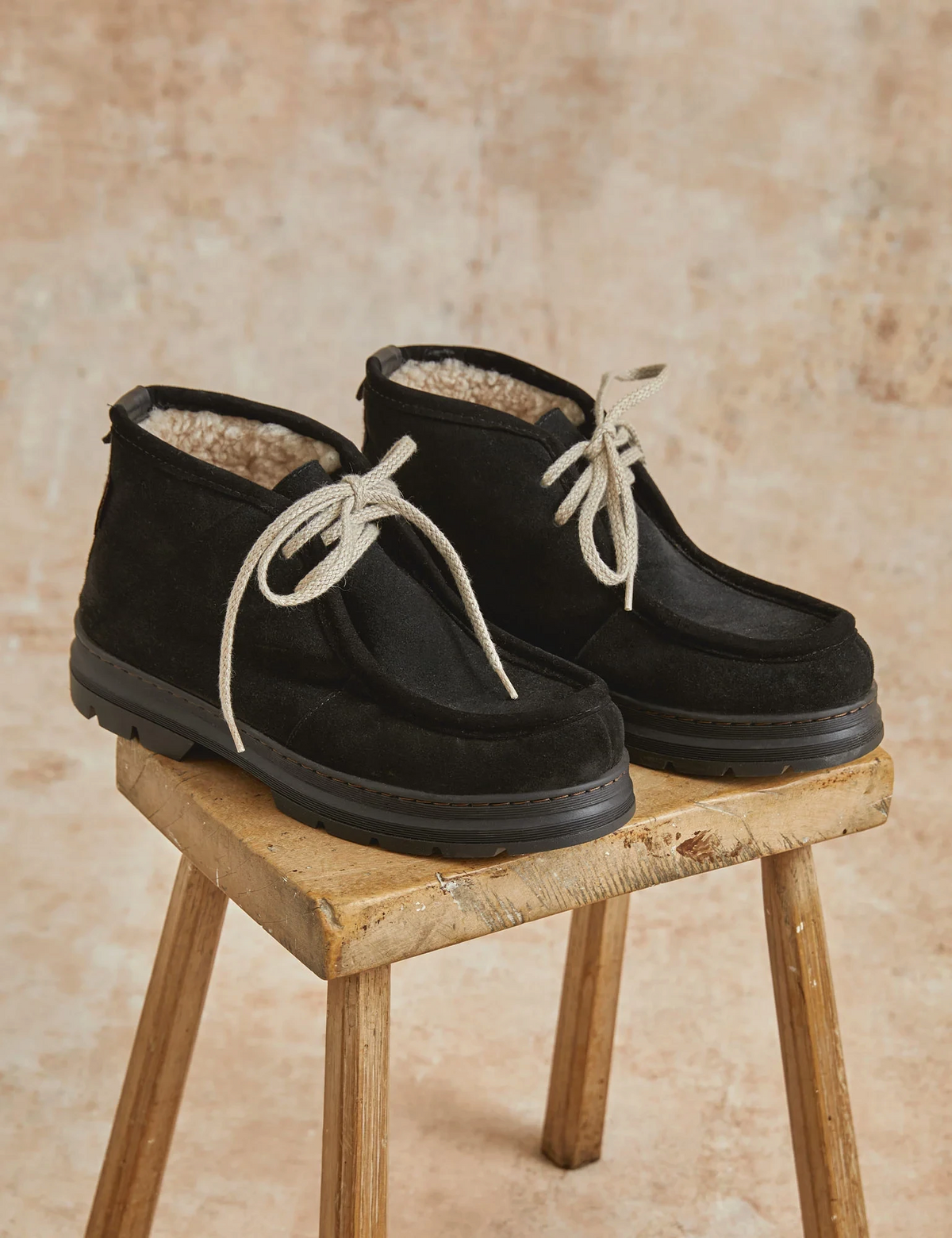 The Copper Shed black split suede shearling-lined boots with ecru laces and a thick rubber tread sole on a stool.
