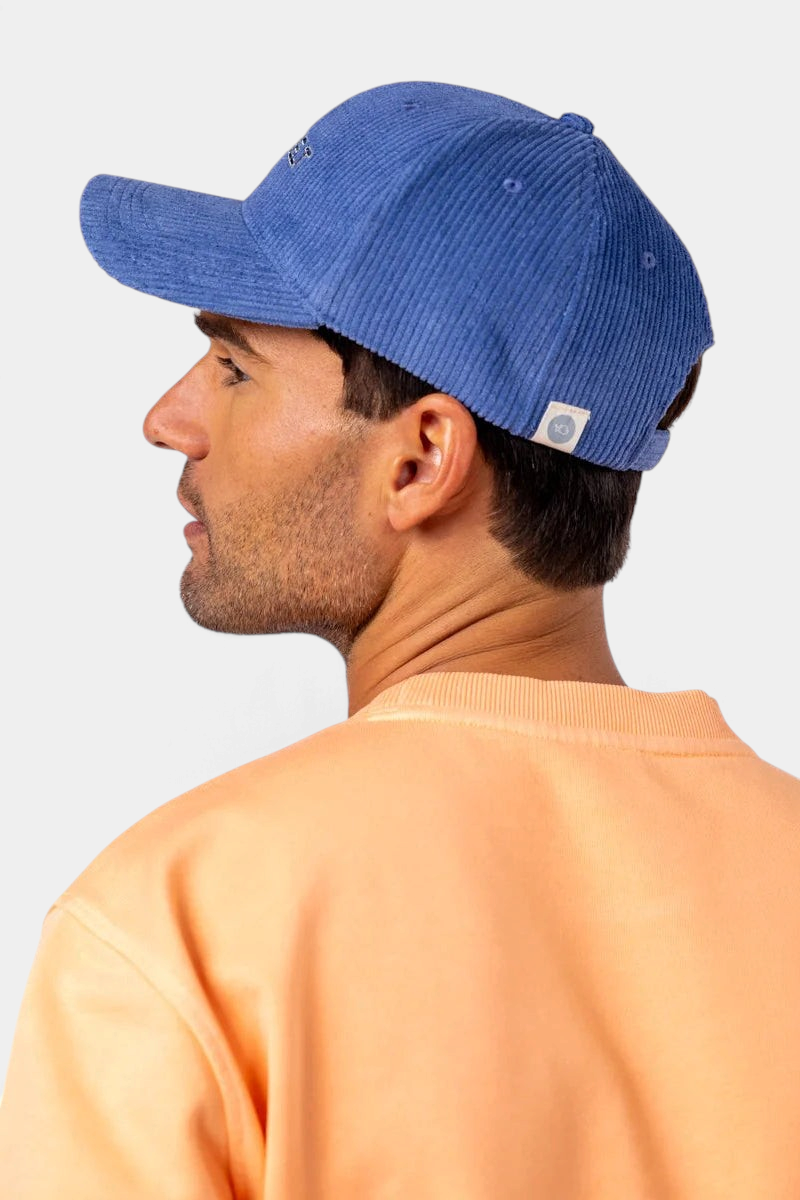 The Copper Shed blue corduroy velvet cap with embroidered logo worn by a man in a peach sweatshirt, side profile view.
