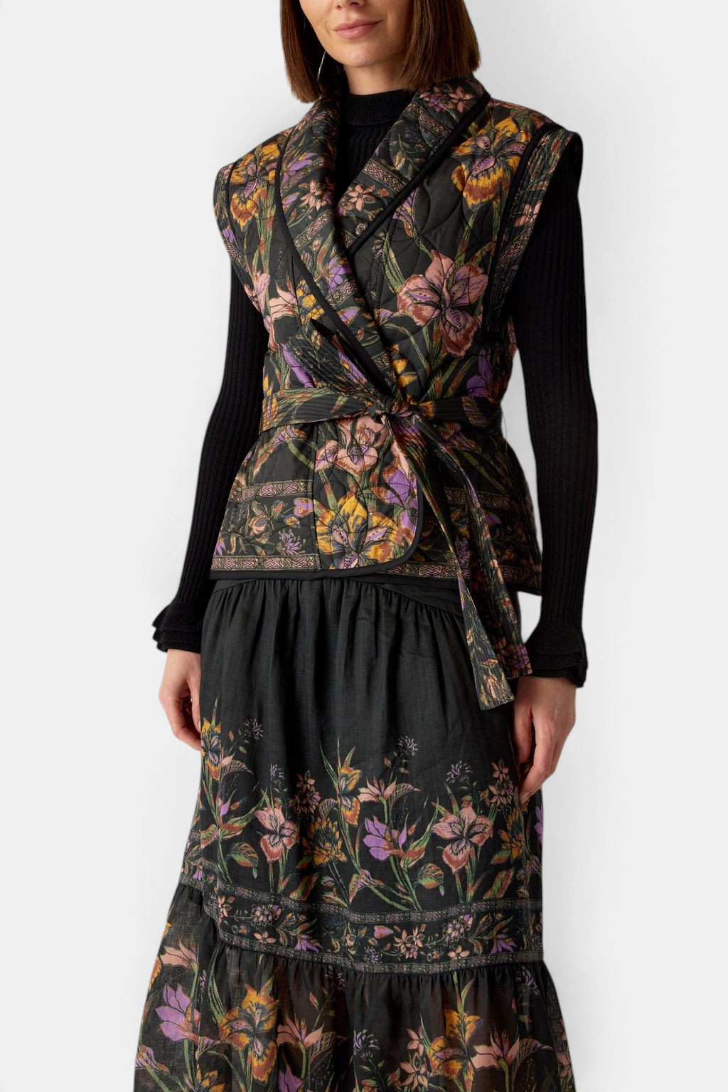 The Copper Shed Bowie vest in a moody floral quilted cotton with a shawl collar and tie belt worn over a black top.