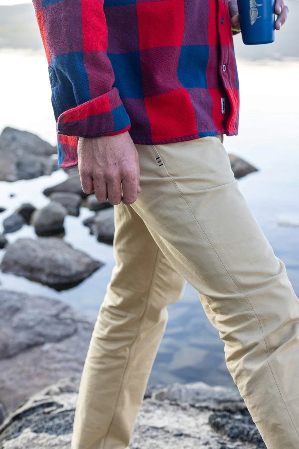 The Copper Shed Breakout broken twill pants in tan, worn by a man standing on rocky ground near a lake.