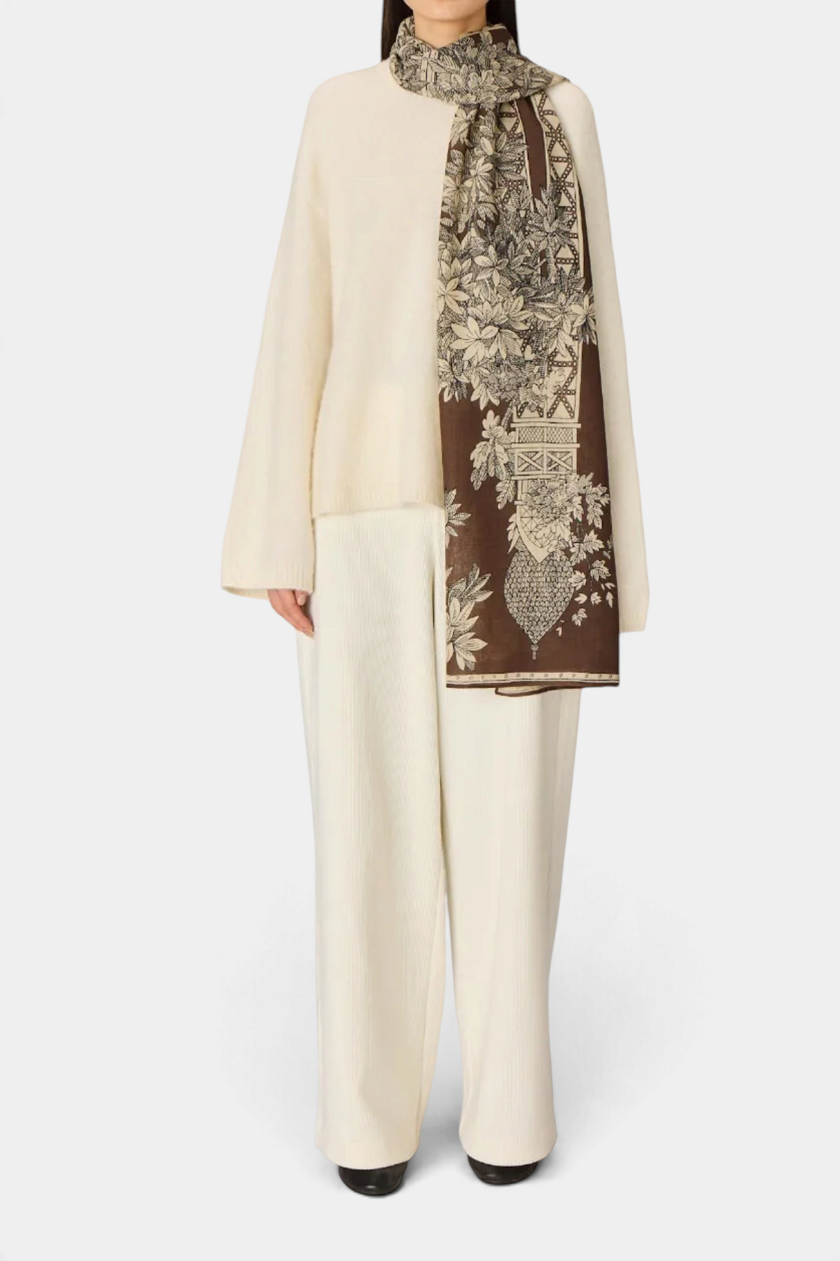 The Copper Shed 100% wool scarf in brown featuring an intricate floral and architectural print worn over cream knitwear