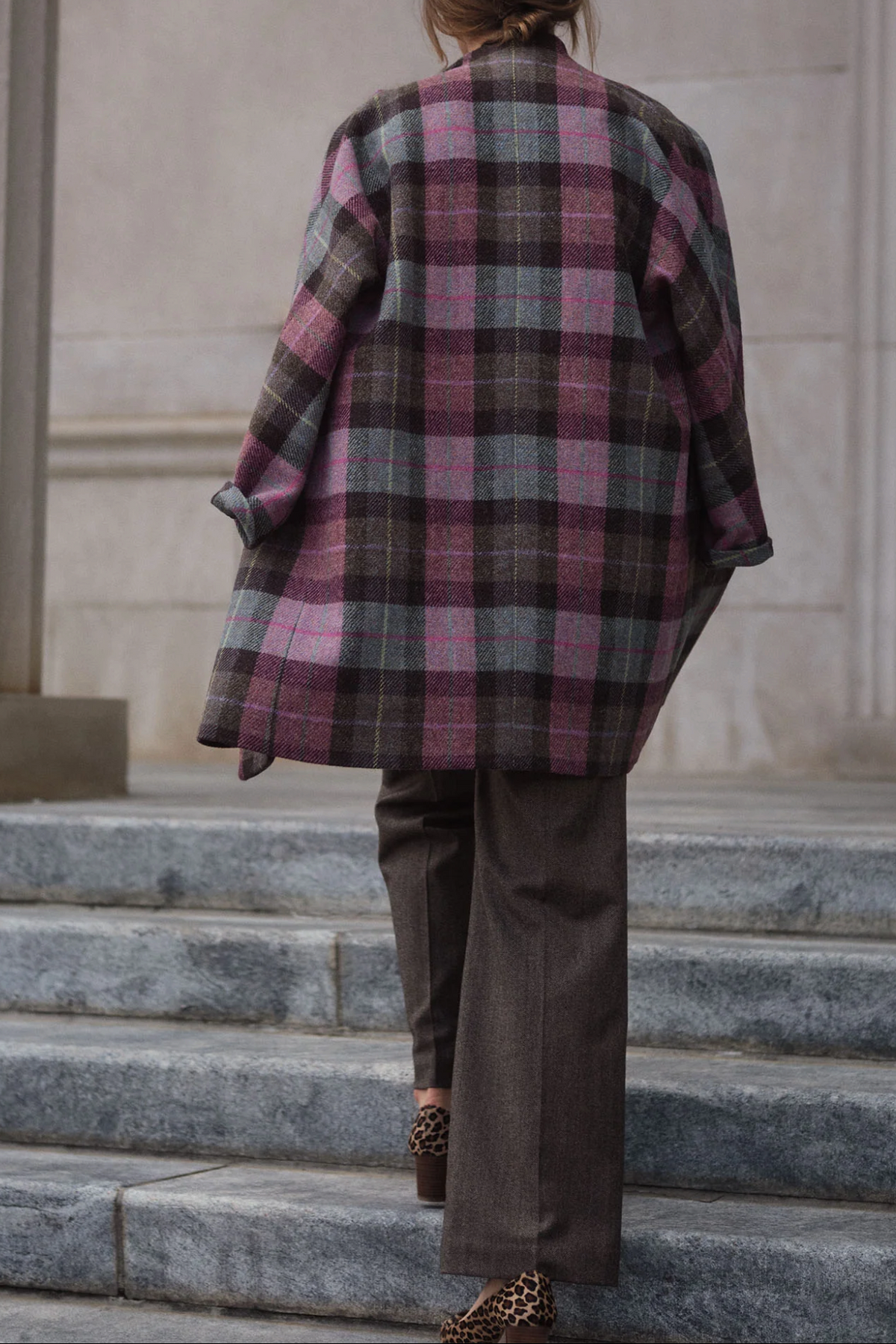 The Copper Shed Kimono Carina coat in a pink and grey Harris Tweed plaid, worn over brown trousers on stone steps.