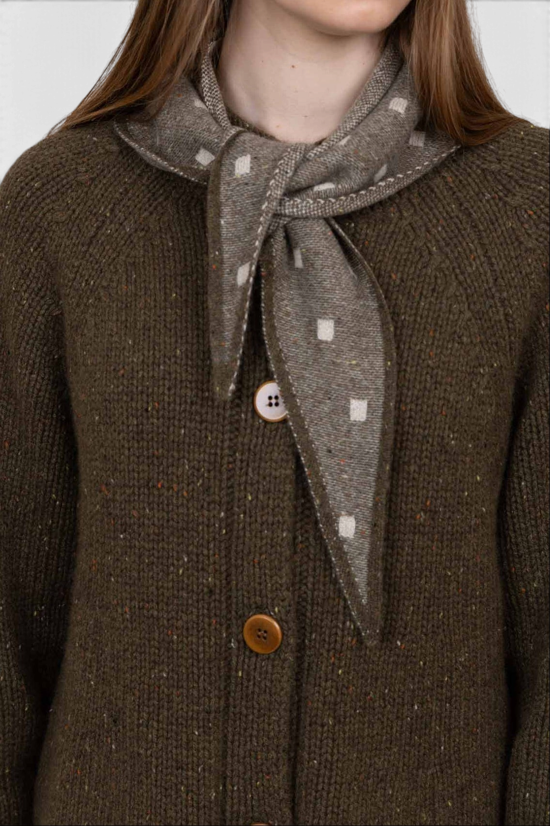 The Copper Shed Mia triangle cashmere neckerchief in taupe with a white square pattern worn over a dark green sweater.