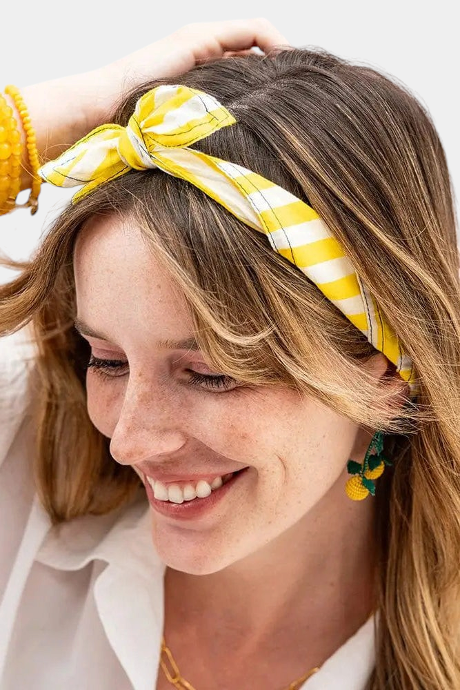 The Copper Shed Naomi yellow and white striped cotton bandana worn as a stylish knotted headband by a smiling woman.
