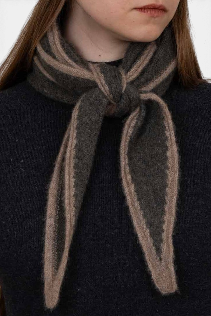 The Copper Shed Austen spindle-shaped knitted organic cashmere scarf in dark grey and beige tied in a French knot.