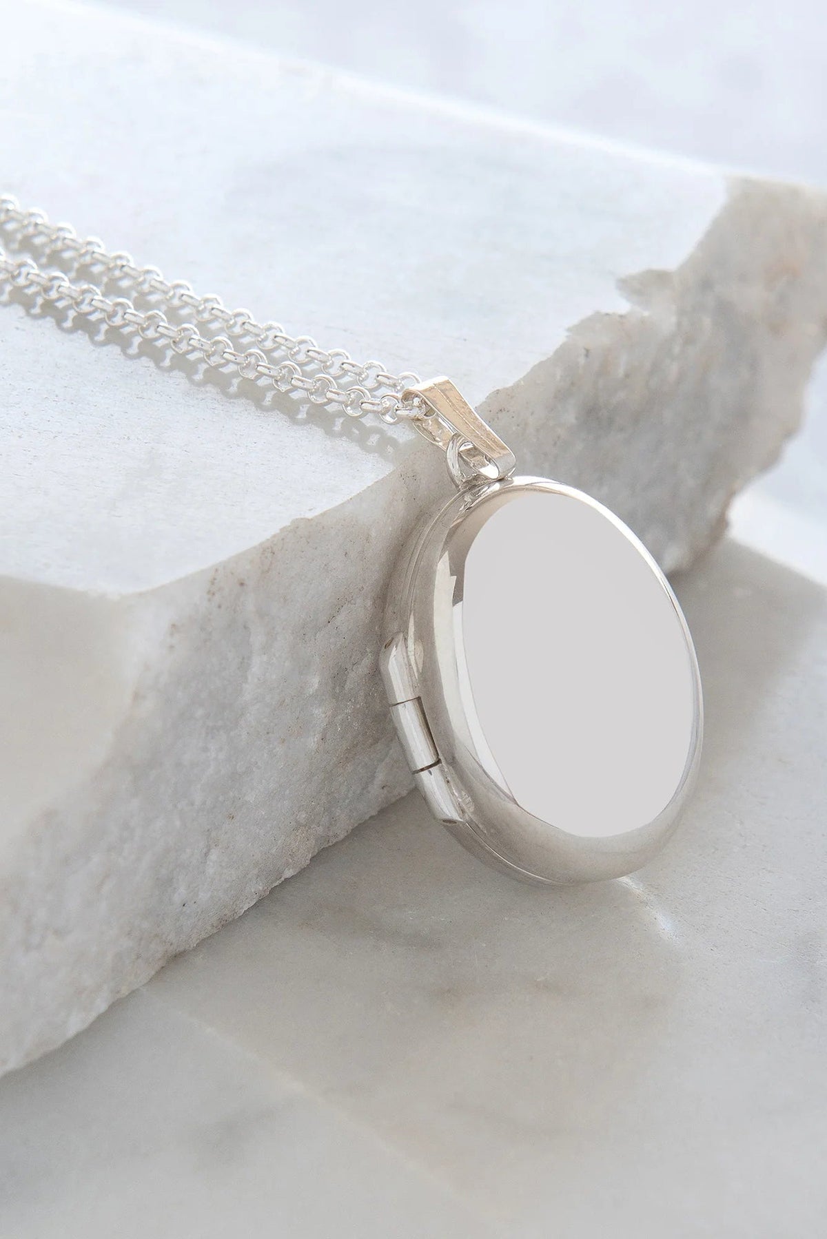 The Copper Shed sterling silver oval locket necklace with a delicate chain resting on a white stone surface.