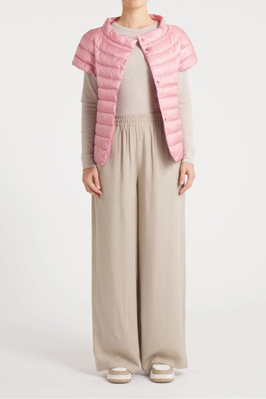 A model stands against a light gray studio background wearing a light pink puffer vest over a beige long-sleeve top and matching beige wide-leg trousers.